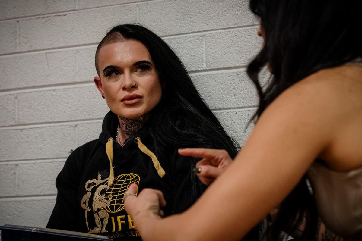Female pro in an IFBB hoodie listening backstage against a cinder block wall
