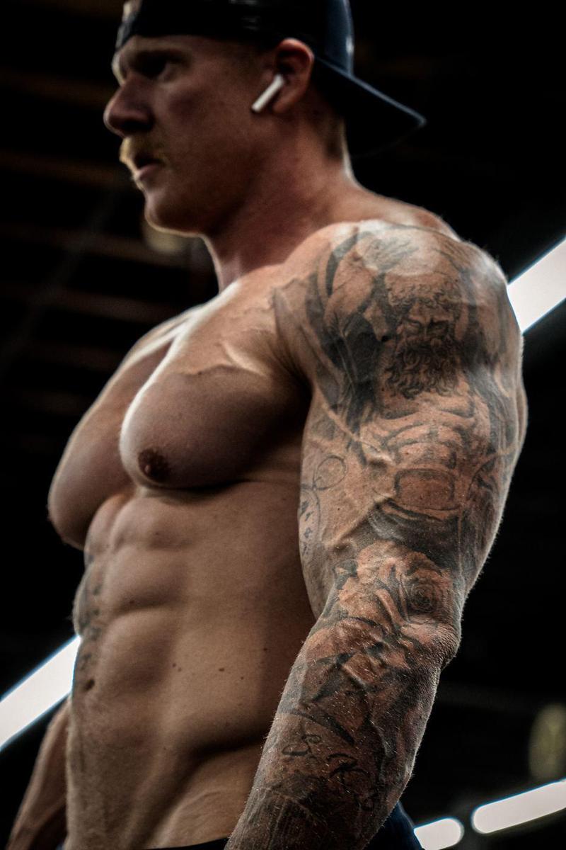 Profile of a tattooed bodybuilder in a backwards cap between sets